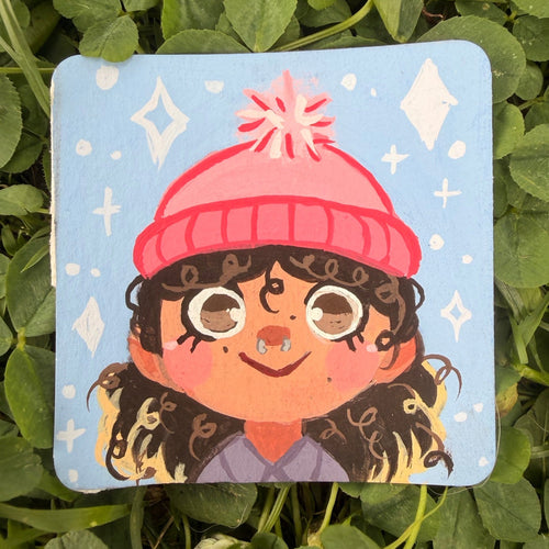 A square painting with rounded corners featuring a cartoon illustration of a girl with curly hair wearing a pink beanie, placed on a bed of green clover.