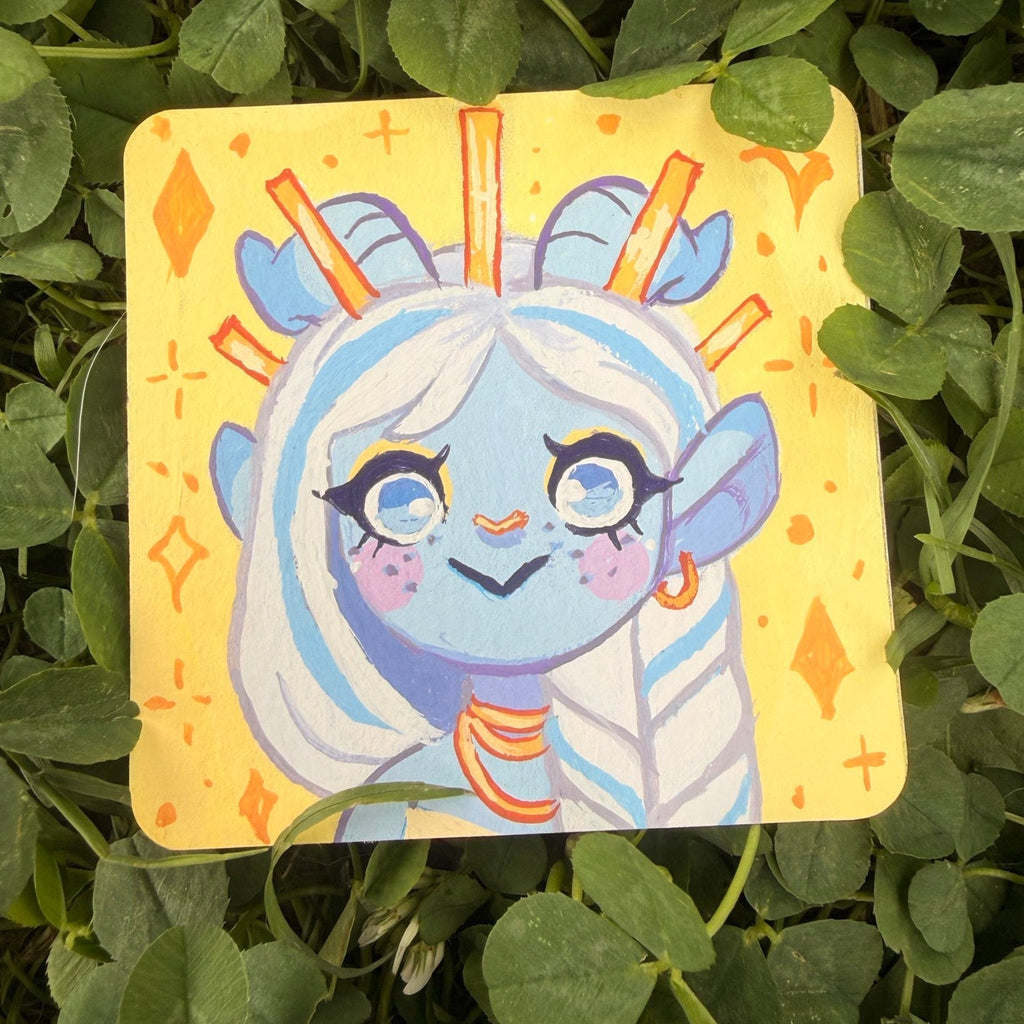 A square yellow card featuring a cute blue cartoon character with horns and a halo placed on green clover leaves outdoors.